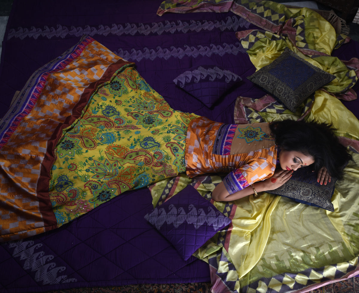 A woman lies on a purple quilted floor surrounded by vibrant yellow, orange, and purple patterned traditional garments and matching pillows.